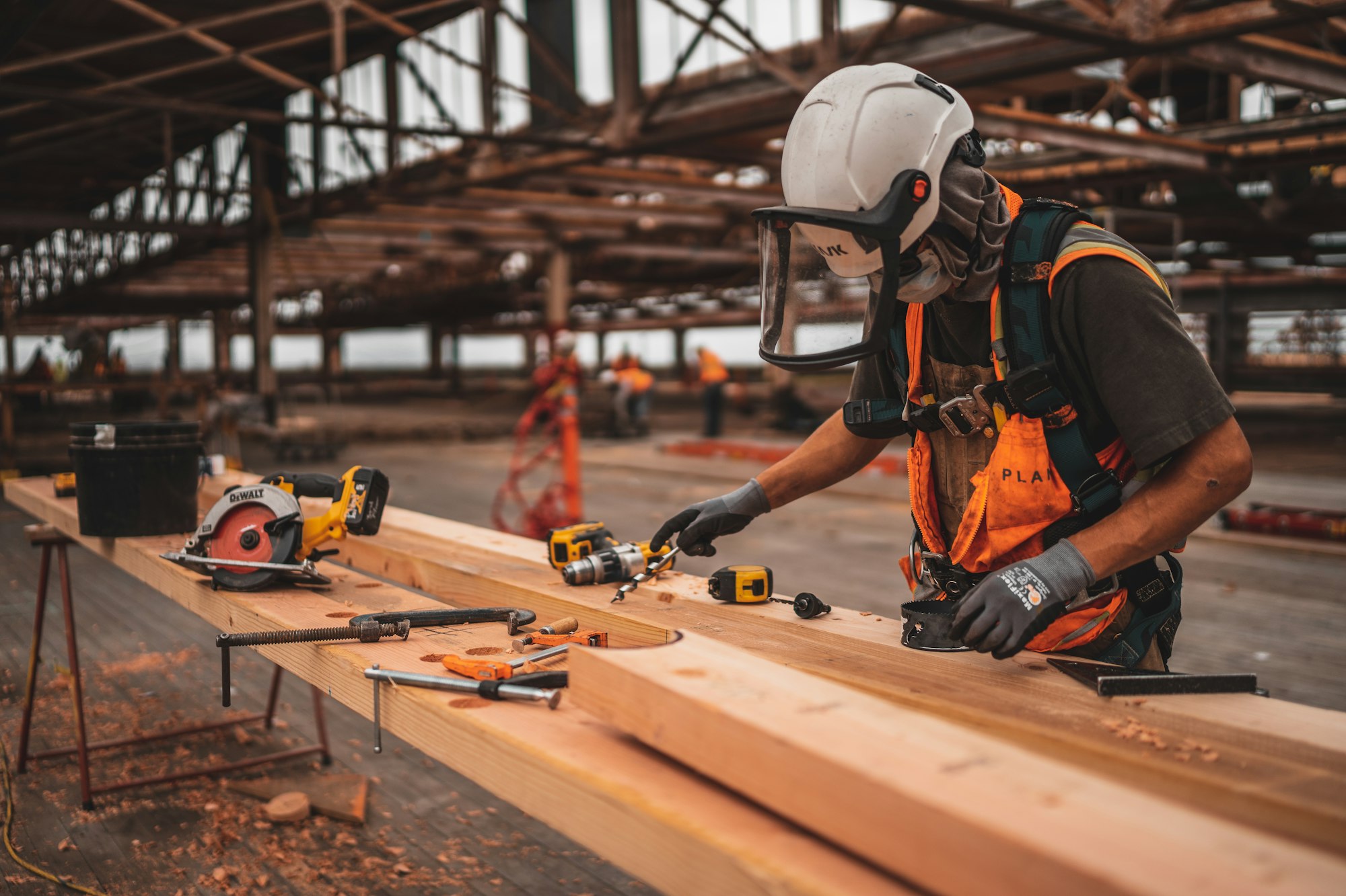 A tradesman in a hard hat and face shield laying out tools on lumber inside a building under construction.